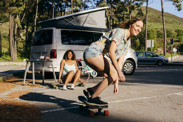 Beautiful skater practicing riding skate board on street with background of woman sitting on skate near minivan in camping - ADSF39352