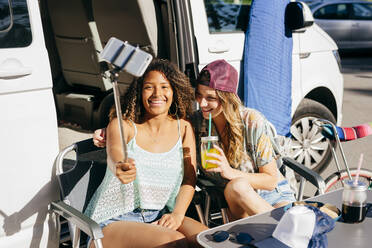 Fröhliche Frauen, die ein Getränk und Nachos genießen, während sie auf dem Campingplatz in einem Van sitzen und ein Selfie mit ihrem Smartphone machen - ADSF39357