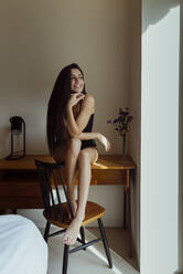 Attraktive, fröhliche junge Frau, die auf einem Tisch sitzt, die Beine auf einen Stuhl legt und in der Nähe eines Fensters in einem Zimmer wegschaut - ADSF39361