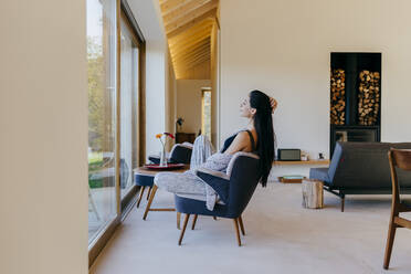 Side view of attractive Hispanic lady sitting on chair in room and looking at big window - ADSF39366