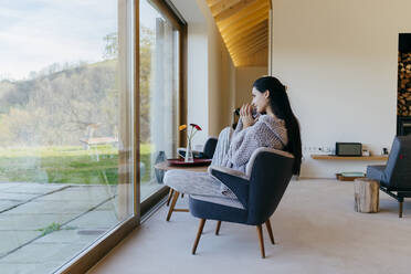 Side view of attractive Hispanic lady sitting on chair in room and looking at big window - ADSF39367