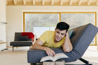 Hispanic happy guy lying on settee and reading a book in room near big window - ADSF39368