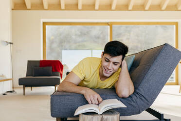 Hispanischer glücklicher Mann, der auf dem Sofa liegt und ein Buch in einem Zimmer in der Nähe eines großen Fensters liest - ADSF39368