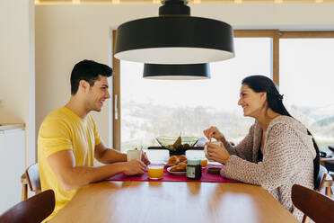 Side view of attractive Hispanic happy lady and young guy having breakfast together at table in modern kitchen - ADSF39370