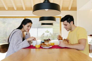 Side view of attractive Hispanic happy lady and young guy having breakfast together at table in modern kitchen - ADSF39373