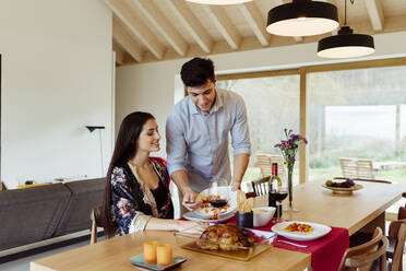 Ein lächelnder Mann serviert einer fröhlichen attraktiven hispanischen Frau am Küchentisch das Essen - ADSF39391