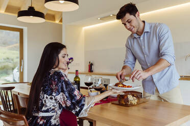 Ein lächelnder Mann serviert einer fröhlichen attraktiven hispanischen Frau am Küchentisch das Essen - ADSF39392