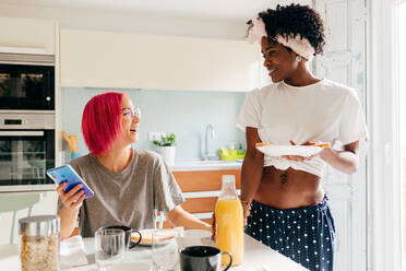 Erfreute junge Frau mit leuchtend rosa Haaren, die lächelt und auf ihrem Smartphone surft, während sie am Tisch in der Nähe eines kochenden afroamerikanischen Freundes beim Frühstück zu Hause sitzt - ADSF39404