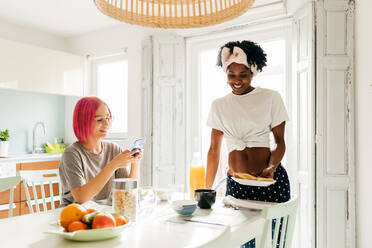Erfreute junge Frau mit leuchtend rosa Haaren, die lächelt und auf ihrem Smartphone surft, während sie am Tisch in der Nähe eines kochenden afroamerikanischen Freundes beim Frühstück zu Hause sitzt - ADSF39405