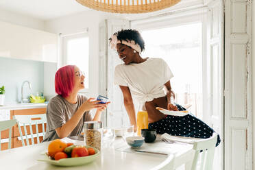 Erfreute junge Frau mit leuchtend rosa Haaren, die lächelt und auf ihrem Smartphone surft, während sie am Tisch in der Nähe eines kochenden afroamerikanischen Freundes beim Frühstück zu Hause sitzt - ADSF39406