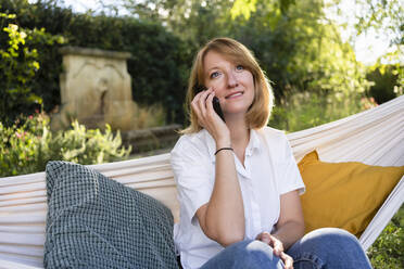 Frau, die in einer Hängematte im Garten sitzt und über ihr Mobiltelefon spricht - SVKF00632
