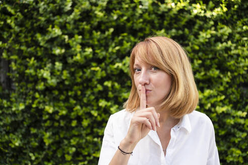 Frau mit Finger auf den Lippen vor einer grünen Hecke stehend - SVKF00640