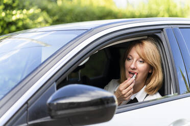 Mature woman looking at side-view mirror of car and applying lipstick - SVKF00642