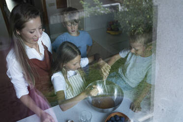 Mutter und Kinder mischen Teig für Kuchen in der Küche, gesehen durch ein Fensterglas - NJAF00038