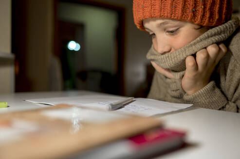 Junge in warmer Kleidung bei den Hausaufgaben am Tisch - ANAF00157