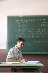 Mathematiklehrer sitzt mit Buch im Klassenzimmer - DAMF01098