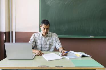 Professor sitzt mit Laptop und überprüft Papier im Klassenzimmer - DAMF01101