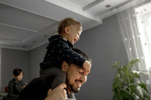 Happy boy sitting on father's shoulder having fun at home - ANAF00162
