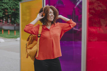 Smiling woman enjoying listening to music on headphones by colorful glasses - YTF00199