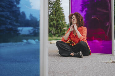Junge Frau mit geschlossenen Augen und Händen am Kinn vor einem magentafarbenen Glas sitzend - YTF00204