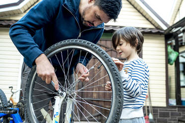 Junge sieht seinen Vater an, der vor dem Haus ein Fahrradrad repariert - ANAF00166