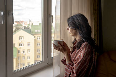 Frau hält Kaffeetasse und schaut durch das Fenster zu Hause - ANAF00174