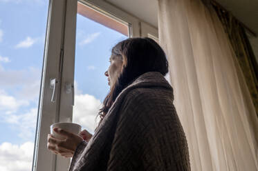 In eine Decke eingewickelte Frau, die eine Kaffeetasse hält und durch ein Fenster zu Hause schaut - ANAF00175