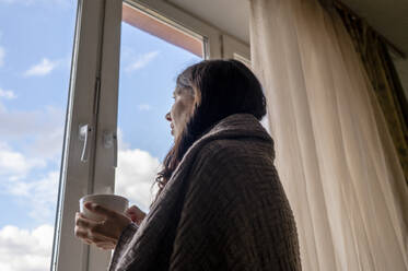 Woman wrapped in blanket holding coffee cup looking through window at home - ANAF00175