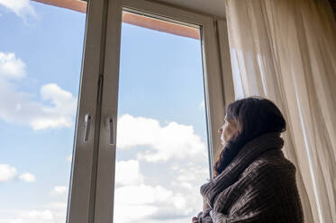 Nachdenkliche, in eine Decke gehüllte Frau, die durch das Fenster ihres Hauses schaut - ANAF00176