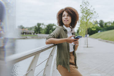 Nachdenkliche junge Frau, die ihr Smartphone hält und sich an ein Geländer lehnt - YTF00209