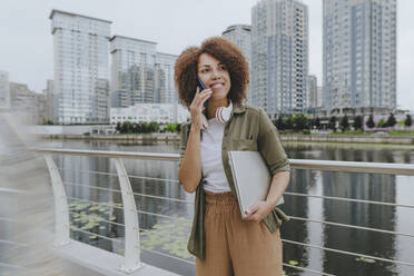 Smiling businesswoman talking on mobile phone standing with laptop in front of river - YTF00212