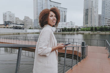 Thoughtful young businesswoman with smart phone standing by railing - YTF00221