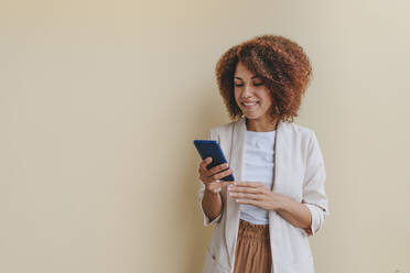 Smiling young businesswoman using mobile phone against beige background - YTF00225