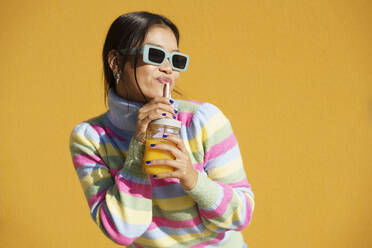 Young woman drinking orange juice standing in front of yellow wall - JSMF02414