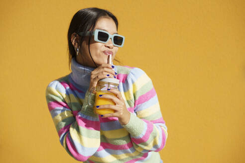Junge Frau trinkt Orangensaft und steht vor einer gelben Wand - JSMF02414
