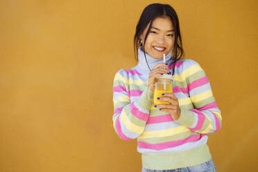 Smiling young woman standing with orange juice in front of wall - JSMF02415