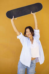 Happy young woman holding aloft skateboard in front of yellow wall - JSMF02420