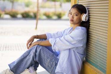 Young woman wearing headphones listening to music leaning on shutter - JSMF02425