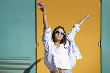 Glückliche junge Frau mit Sonnenbrille, die sich an einem sonnigen Tag vor einer Mauer amüsiert - JSMF02426
