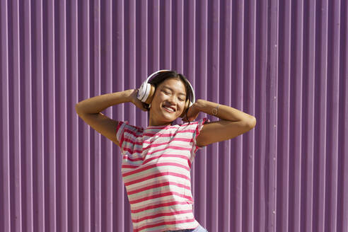 Smiling young woman wearing headphones listening music in front of purple wall - JSMF02437
