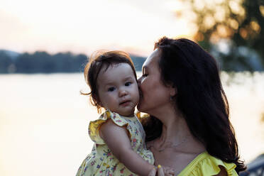 Mother kissing daughter in front of river at sunset - JBUF00013