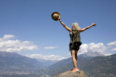 Carefree senior woman with arms raised standing on rock in front of mountains - FLLF00785