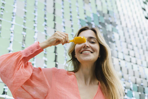 Happy woman covering eye with ice cream in front of glass wall - SEAF01315