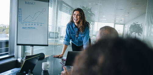 Smiling businesswoman standing at table and discussing finance with team. Businesswoman giving presentation over a project budget to colleagues in office boardroom. - JLPSF05198