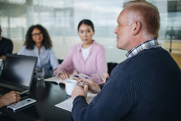 Senior Geschäftsmann, der eine Besprechung mit seinem Team abhält. Geschäftsleute, die im Sitzungssaal des Büros sitzen und ein Projekt besprechen. - JLPSF05204