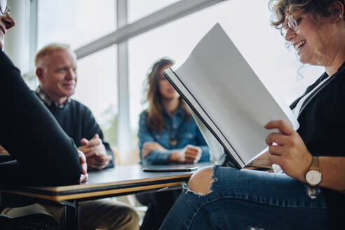 Lächelnde Geschäftsfrau, die ein Papier mit ihren Kollegen liest, die im Besprechungsraum sitzen. Weibliche Fachkraft, die ein Vertragsdokument während einer Besprechung im Sitzungssaal liest. - JLPSF05220