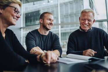 Geschäftsmann, der Papierkram betrachtet und lächelt, während er in einer Teamsitzung im Sitzungssaal sitzt. Gruppe von glücklichen Geschäftsleuten während einer Sitzung im Konferenzraum. - JLPSF05224