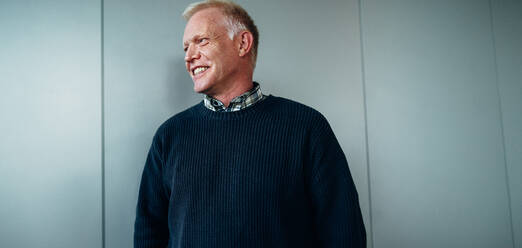 Smiling businessman standing against a wall. Senior entrepreneur wearing sweater looking away and smiling. - JLPSF05243