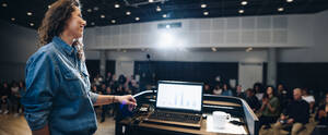 Weitwinkelaufnahme einer Rednerin, die auf einem Podium steht, mit einem Laptop auf dem Rednerpult und Menschen, die im Auditorium sitzen. Eine Geschäftsfrau, die eine Präsentation bei einer Firmenveranstaltung hält. - JLPSF05269