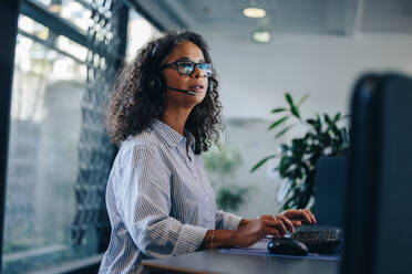 Eine Frau arbeitet am Computer und spricht über ein Headset. Eine Geschäftsfrau mit Headset arbeitet an ihrem Schreibtisch. - JLPSF05280